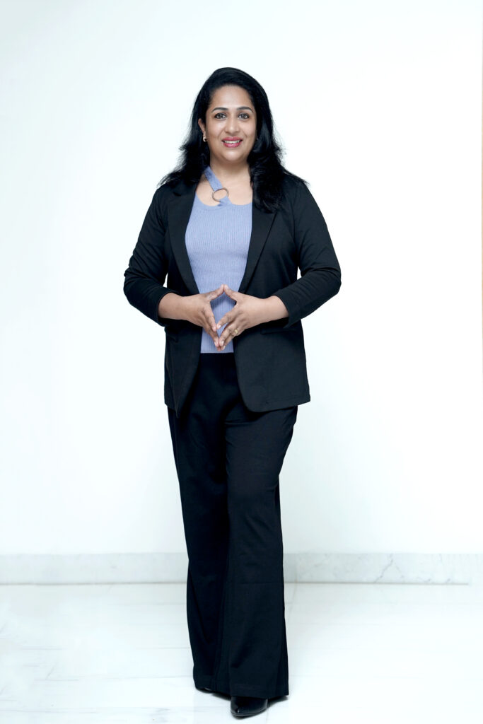 A smiling Indian counseling psychologist and NLP practitioner, Radhikaa Riju Nairr is shown in this full-length portrait standing confidently against a white background with her hands clasped. She is dressed in a black blazer and light purple pants.