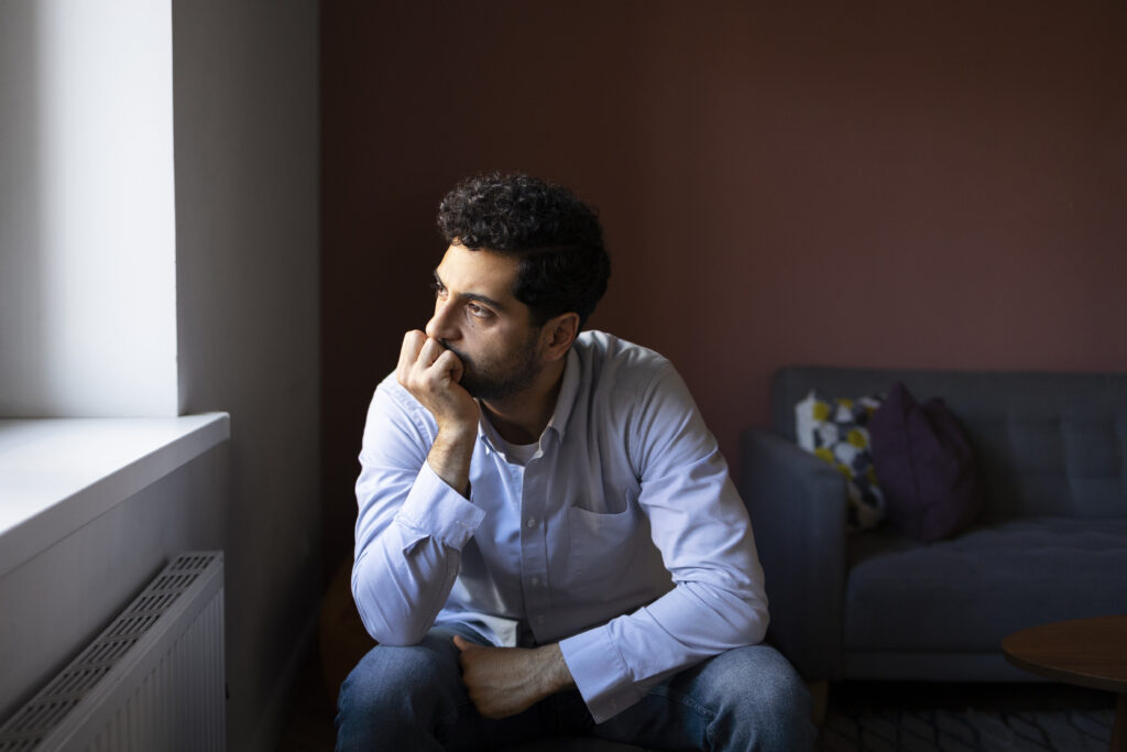 Young man sitting by a window, looking thoughtful and anxious, representing emotional stress or anxiety issues.