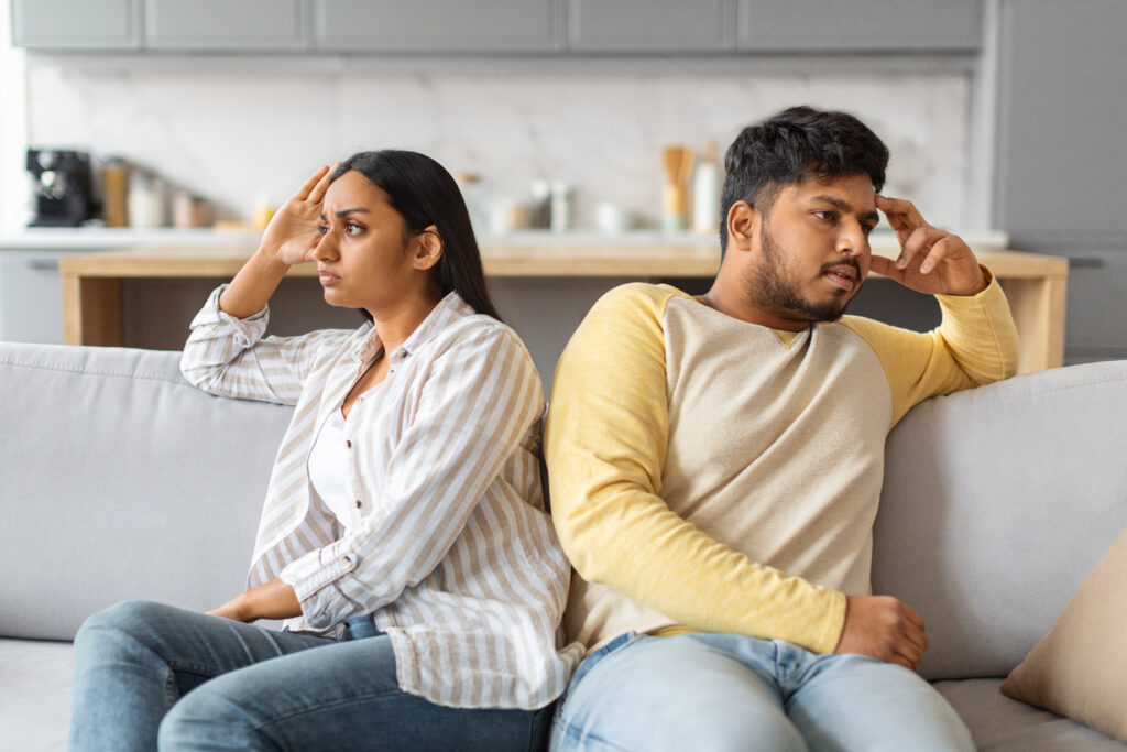 Unhappy couple sitting apart on a couch, both looking upset and disconnected, symbolizing communication breakdown in relationships.